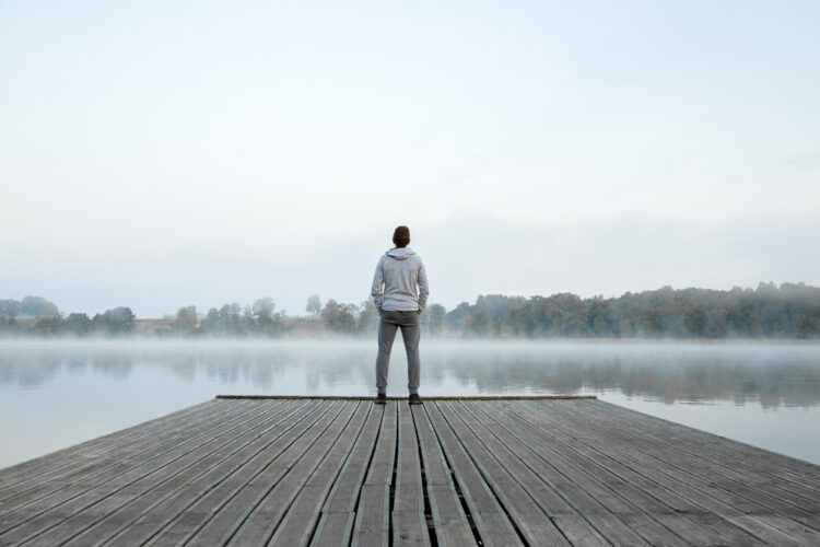 Young man standing alone on wooden footbridge and staring at lake. Thinking about life. Mist over water. Foggy air. Early chilly morning. Peaceful atmosphere in nature. Enjoying fresh air. Back view.