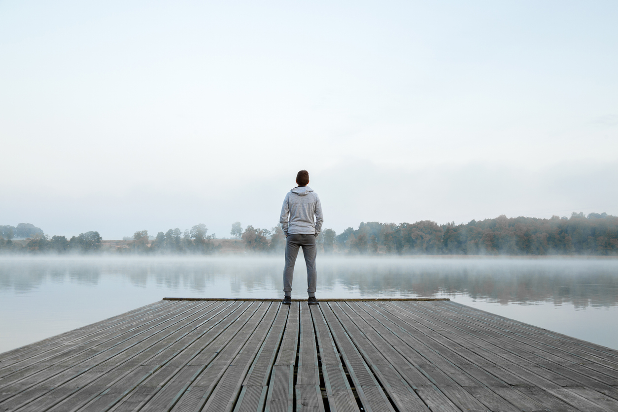 Young man standing alone on wooden footbridge and staring at lake. Thinking about life. Mist over water. Foggy air. Early chilly morning. Peaceful atmosphere in nature. Enjoying fresh air. Back view.