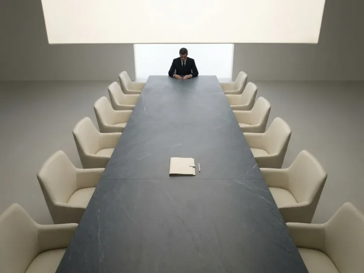Persoon alleen aan het hoofd van een lange minimalistische vergadertafel, omringd door lege stoelen, met één document in het midden.