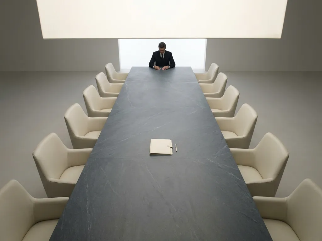 Persoon alleen aan het hoofd van een lange minimalistische vergadertafel, omringd door lege stoelen, met één document in het midden.