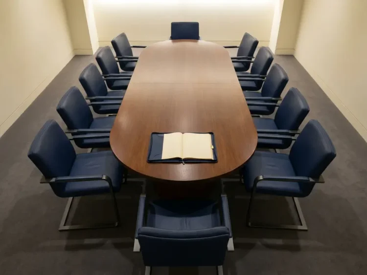 Ovale vergadertafel van bovenaf met lege leren stoelen en een opengeslagen dossier in het midden, minimalistische boardroom.