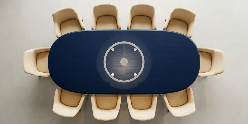 vergaderzaal-ovale-tafel-bovenaanzicht-symmetrie.jpg Ovale vergadertafel van bovenaf met acht symmetrisch geplaatste stoelen in crème en marineblauw tinten, verlicht door één spotlight.